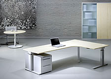 H&ouml;henverstellbarer Schreibtisch Spine&sup2;: Eckschreibtisch 240 x 160 x 80 cm mit Oberfl&auml;che in Ahorn-Dekor und kristallwei&szlig;em Gestell. Im Hintergrund runder Besprechungstisch.