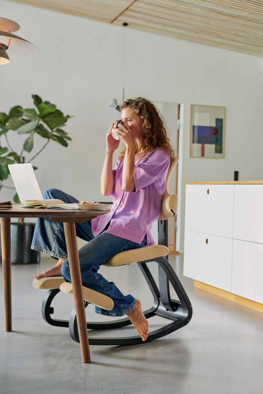 Frau trinkt Kaffee in alternativer Sitzhaltung auf dem Stokke Thatsit mit einem Fuß auf dem Kniepolster